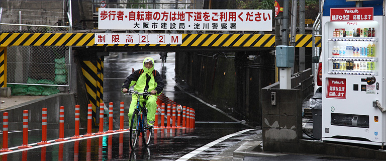 Fahrradfahrer im verregneten Osaka, rechts einer der in Japan sehr verbreiteten Getr&auml;nkeautomaten.